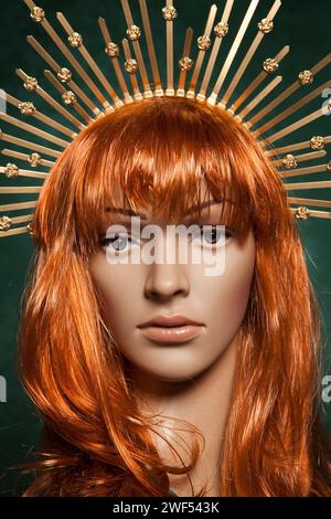 Mannequin de femme aux cheveux rouges en plastique avec de longs cheveux brillants portant une couronne dorée piquante posant sur un fond vert Banque D'Images