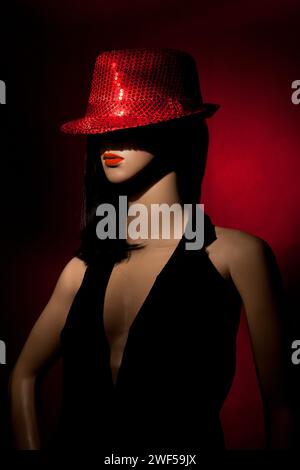 Mannequin de femme en plastique avec cheveux longs clack, rouge à lèvres rouge, et portant un chapeau de paillettes rouge vif posant sur un fond rouge dans un set studio d'une lumière Banque D'Images