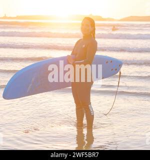 Surfeuse souriante en combinaison humide tient sa planche de surf au coucher du soleil à Essaouira, Maroc, le 28 janvier 2024 Banque D'Images