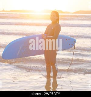 Surfeuse souriante en combinaison humide tient sa planche de surf au coucher du soleil à Essaouira, Maroc, le 28 janvier 2024 Banque D'Images