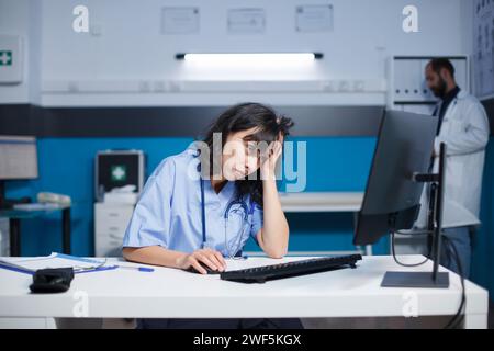 Femme caucasienne dans Blue Scrubs, étudiant lassablement les données médicales sur son ordinateur de bureau. Dans un hôpital, une infirmière à l'air fatiguée utilise un ordinateur tandis qu'un homme en blouse de laboratoire travaille en arrière-plan. Banque D'Images