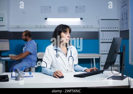 Dans le bureau de la clinique, un jeune médecin caucasien engagé examine les dossiers médicaux sur un ordinateur. La praticienne vérifie les informations du patient sur un ordinateur de bureau à l'hôpital. Banque D'Images