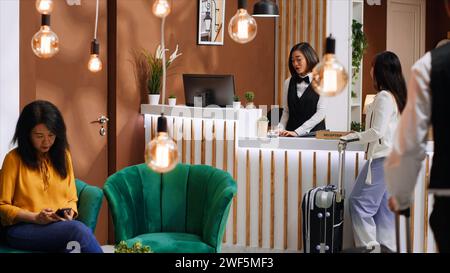 Jeune voyageur arrivant dans le hall de l'hôtel avec des bagages, réceptionniste demandant à la cliente de réserver sa chambre pour l'enregistrement. Employé accueillant le client au complexe cinq étoiles, industrie hôtelière. Banque D'Images