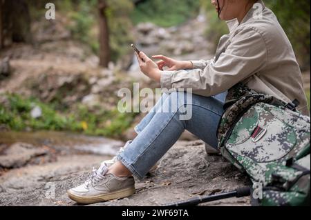 Une image vue de côté d'une voyageuse de trekker asiatique positive est assise sur un rocher et utilise son smartphone, prenant une pause tout en randonnant sur la montagne Banque D'Images