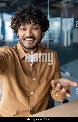 Vue à la première personne d'un homme musulman dans des vêtements décontractés intelligents ayant un appel vidéo téléphonique et souriant tout en étant assis au bureau. Manager inspiré montrant à la famille un nouvel espace de travail utilisant une connexion Internet sans fil. Banque D'Images