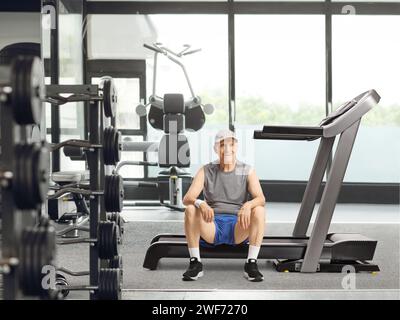 Homme âgé assis sur un tapis roulant dans une salle de gym et souriant à la caméra Banque D'Images