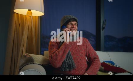 Heureux jeune homme porter une écharpe et un bonnet parlant avec un téléphone portable tout en étant assis sur le canapé dans le salon. Homme relaxant à la maison, communiquant avec famil Banque D'Images