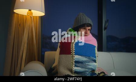 Jeune homme porter une écharpe et un bonnet couvrant la couverture lui-même pour le froid tout en étant assis sur le canapé dans le salon. L'homme exprime un sentiment de tristesse et de discomfo Banque D'Images