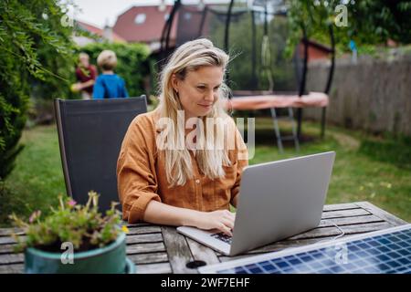 Femme d'affaires, freelance ou gestionnaire travaillant à l'extérieur dans le jardin, jardin homeoffice. Travail à distance depuis l'arrière-cour. L'ordinateur portable se charge avec le chargeur solaire Banque D'Images