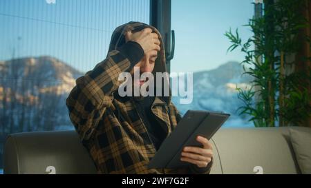 Jeune homme barbu porter un sweat à capuche réagissant à la perte, aux mauvaises nouvelles tout en utilisant une tablette. L'homme est contrarié, ressent de l'anxiété et du stress assis sur le canapé en livi Banque D'Images