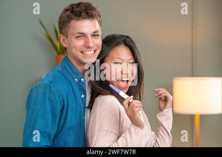 Couple multiracial excité avec test de grossesse positif regardant la caméra. Intérieur Banque D'Images