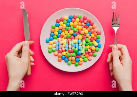 la fille tient des couverts dans ses mains et mange des bonbons dans une assiette. Concept de santé et d'obésité, vue de dessus sur fond coloré. Banque D'Images