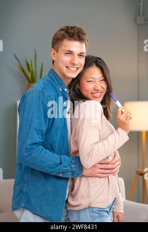 Portrait vertical excité couple multiracial avec test de grossesse positif regardant la caméra. Intérieur Banque D'Images