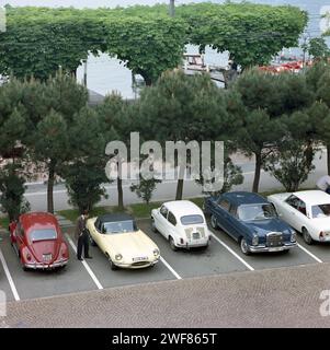 Un spectateur inspecte une Jaguar E-type de 1967, cabriolet, Pale Primrose Yellow, S1 1/2 stationnée sur le front de mer du lac Léman à Lausanne, à côté d'autres voitures. Banque D'Images