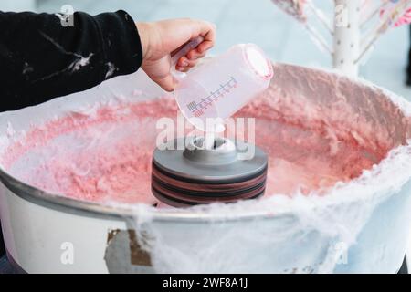 Gros plan d'une main recadrée méconnaissable versant du sucre dans une machine à bonbons de coton avec du fil rose à bonbons se formant à l'intérieur Banque D'Images