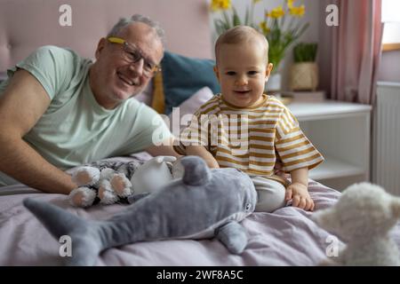 Un grand-père joyeux allongé à côté d'un enfant heureux, entouré de jouets en peluche dans une pièce confortable Banque D'Images