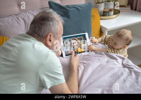 Un homme âgé et un bébé passent un appel vidéo avec une jeune femme sur une tablette numérique tout en étant confortablement assis sur un lit orné d'oreillers Banque D'Images