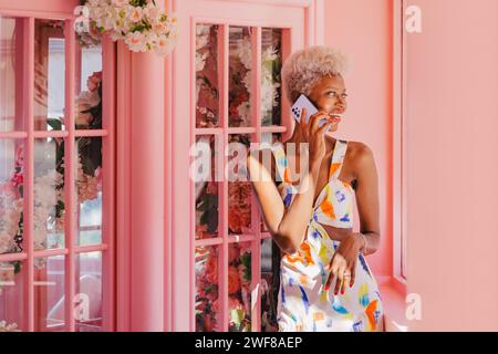 Positive jeune femme panaméenne en tenue élégante souriant et parlant sur téléphone portable tout en se tenant près de la fenêtre rose dans la chambre confortable Banque D'Images