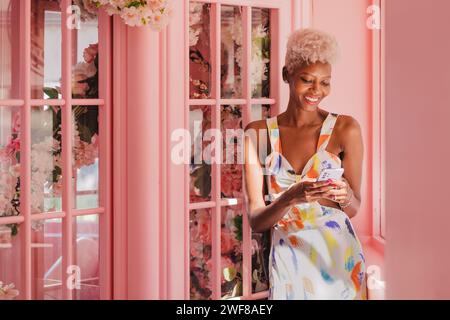 Positive jeune femme panaméenne en tenue élégante souriant et regardant l'écran du smartphone tout en se tenant près de la fenêtre rose dans la chambre confortable Banque D'Images