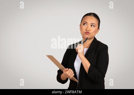 Pensive femme d'affaires asiatique tenant un presse-papiers et un stylo à son menton, l'air réfléchi et concentré Banque D'Images