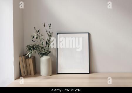 Élégant espace de travail méditerranéen. Livres, maquette de cadre noir vertical vide. Fond de mur blanc. Bureau en bois, table. Vase avec olive Banque D'Images