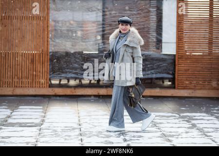 Femme mature élégante marche sur toute la longueur dans la rue en hiver, porte une tenue multicouche à la mode, manteau veste en laine grise, costume avec pantalon et col roulé Banque D'Images