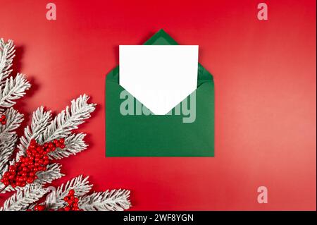 Vue de dessus de l'enveloppe verte, carte blanche, baies rouges, branches de sapin sur fond rouge. Noël, composition du nouvel an. Banque D'Images