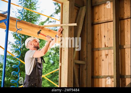 Menuisier construisant un cadre en bois, maison de deux étages près de la forêt. Homme barbu martelant des clous avec un marteau tout en habillant une combinaison de travail. Concept de construction moderne respectueuse de l'environnement. Banque D'Images