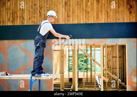 Carpenter construisant une maison en bois encadrée. Homme ouvrier en lunettes habillage façade de maison avec panneaux de particules de ciment, les fixant avec des tournevis. Concept de construction écologique moderne. Banque D'Images