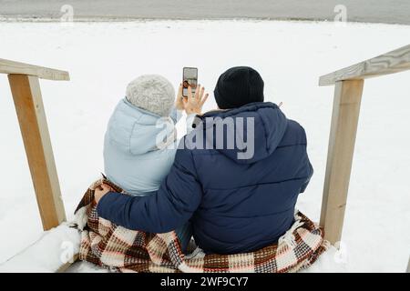 Heureux couple de personnes âgées assis ensemble sur les escaliers au bord de la mer en hiver, prenant un selfie. Banque D'Images