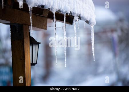 Glaçons de neige suspendus à un toit de bâtiments en bois. Une lanterne en arrière-plan et des arbres enneigés. Banque D'Images