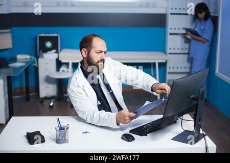 L'image montre un médecin engagé examinant et analysant ses notes au bureau de la clinique. Jeune homme se préparant pour les consultations médicales des patients tout en utilisant un ordinateur de bureau et un presse-papiers. Banque D'Images