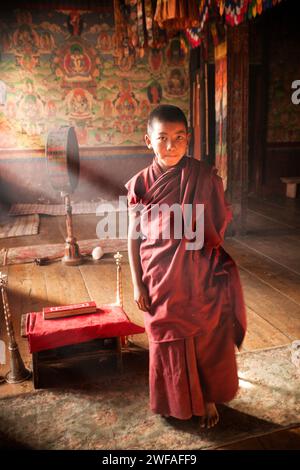 Un jeune moine pieds nus dans une robe rouge se tient sur un tapis altéré au-dessus d'un plancher de bois à l'intérieur de l'ancien temple bouddhiste où il réside dans le village d'Ura Banque D'Images