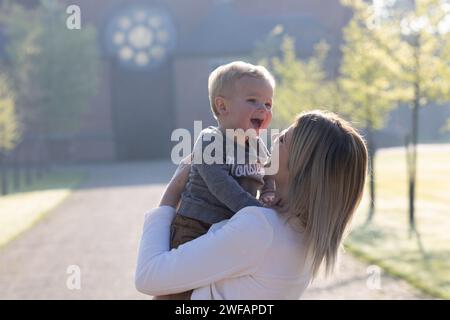 Cette charmante capture montre une jeune femme caucasienne soulevant un enfant en bas âge dans les airs, les deux se prélassant dans l'éclat d'une matinée joyeuse. Alors que le tout-petit rit de bon cœur, ses yeux plissant de joie, la femme le regarde avec une expression d'amour et de bonheur. La lumière ambiante du début de journée les entoure, renforçant l'énergie exubérante et aimante de leur interaction. Gleeful Mornings : une Symphonie du rire. Photo de haute qualité Banque D'Images