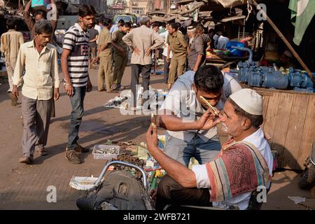 Un coiffeur en plein air rase un client à Chor Bazar (marché des voleurs), Mumbai, en Inde, tandis que ce dernier observe les progrès dans un petit miroir à main Banque D'Images