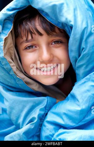 Portrait de garçon, d'enfant et de tente pour le camping, la détente et les vacances d'été heureuses dans les voyages en plein air. Jeune campeur, sourire et enveloppé dans un sac de couchage Banque D'Images