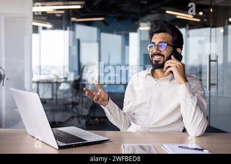 Jeune homme d'affaires indien prospère assis dans le bureau au bureau et parlant au téléphone avec un sourire. Banque D'Images