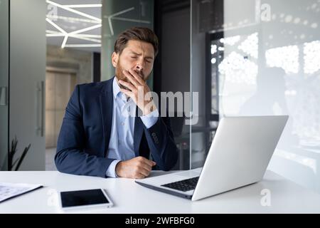 Jeune homme d'affaires fatigué dans un costume assis dans le bureau au bureau et bâille, couvre sa bouche avec sa main, se sent endormi. Banque D'Images