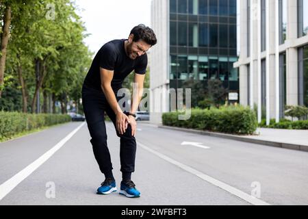 Un jeune homme coureur et sportif se tient dans la rue de la ville en vêtements de sport et tient sa main par sa jambe, ressent une douleur intense au genou, grimacé de la blessure après l'entraînement. Banque D'Images