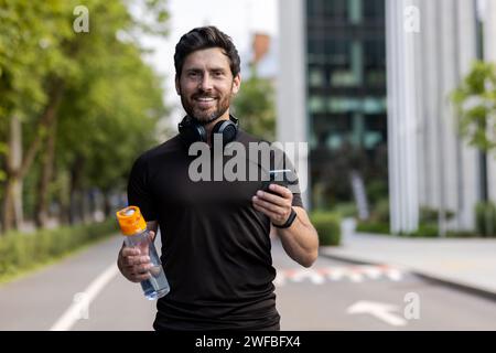 Portrait d'un jeune homme souriant courant dans la rue de la ville, debout dans un casque, tenant une bouteille d'eau et un téléphone, regardant la caméra. Banque D'Images