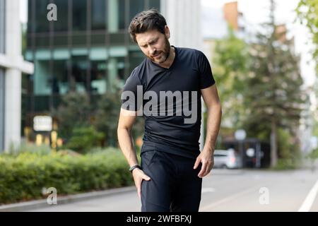 Un jeune homme coureur se tient dehors dans la rue et tient sa jambe, tire un muscle, donne un massage, souffre de douleurs. Banque D'Images