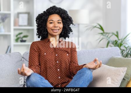 Portrait d'une jeune femme afro-américaine souriante assise sur le canapé à la maison dans la position du lotus de yoga et regardant la caméra. Gros plan. Banque D'Images