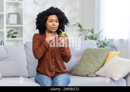 Jeune femme afro-américaine assise sur un canapé à la maison, tenant sa gorge, souffrant de douleurs et utilisant un spray médical. Banque D'Images