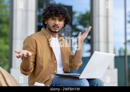 Portrait d'un Indien déçu et inquiet assis avec un ordinateur portable sur un banc, regardant la caméra, écartant ses mains. Banque D'Images
