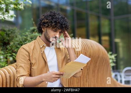 Photo en gros plan d'un jeune Indien assis sur un banc dehors et lisant tristement une lettre dans une enveloppe, tenant avec inquiétude sa tête après avoir reçu de mauvaises nouvelles. Banque D'Images