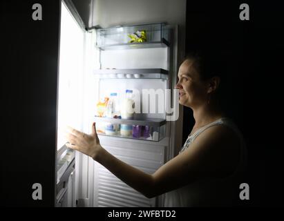 Une femme souriante la nuit à la maison a ouvert le réfrigérateur Banque D'Images