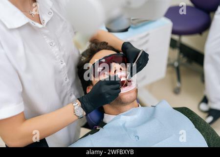 Gros plan d'une dentiste méconnaissable utilisant le laser dentaire à diode moderne pour les soins parodontaux. Patient masculin portant des lunettes de protection, prévention Banque D'Images