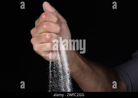 Homme avec une poignée de sable blanc sec dans ses mains, répande du sable à travers les doigts sur fond noir. Concept de flux de vie. Gros plan de grains de pur Banque D'Images