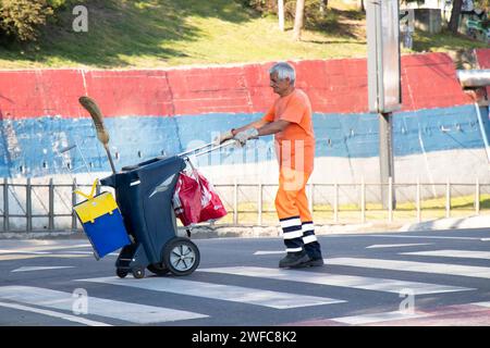 Belgrade, Serbie - 5 juin 2021 : le collecteur d'ordures du service public de la ville traverse la rue tout en poussant le chariot de poubelle de taille avec balai et dustbi Banque D'Images