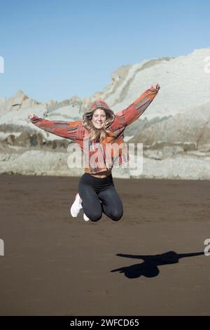 femme saute joyeusement sur une plage, projetant une ombre, avec des falaises rocheuses en arrière-plan Banque D'Images